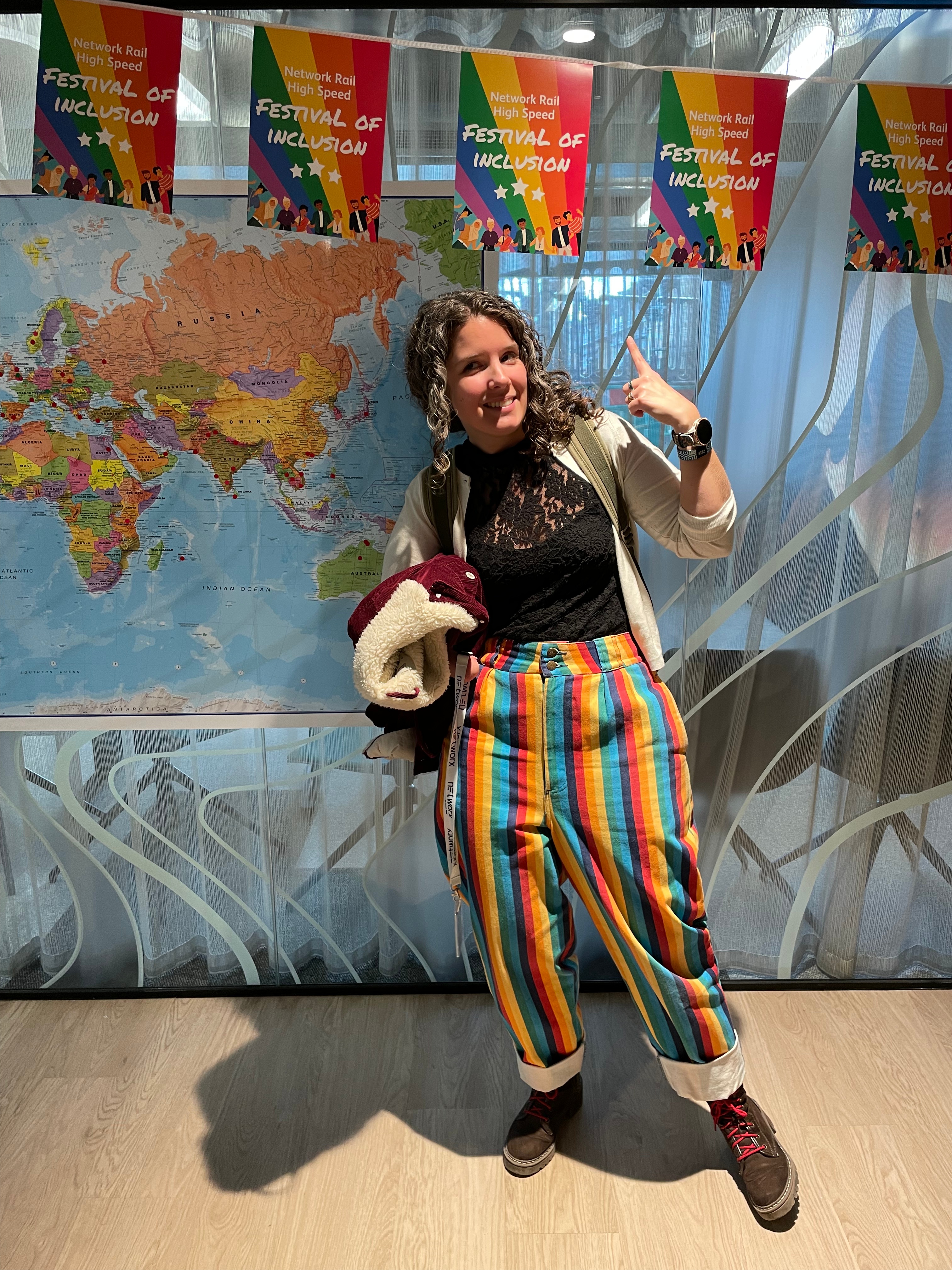 Elisabeth in rainbow trousers pointing at a sign saying Network Rail high speed Festival of inclusion bunting after delivering a talk.
