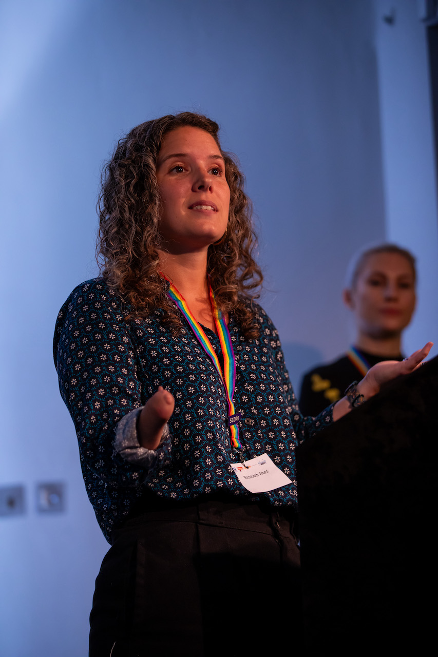 Elisabeth delivering a speech, she has brown curly hair and is gesturing. She's missing her right hand.